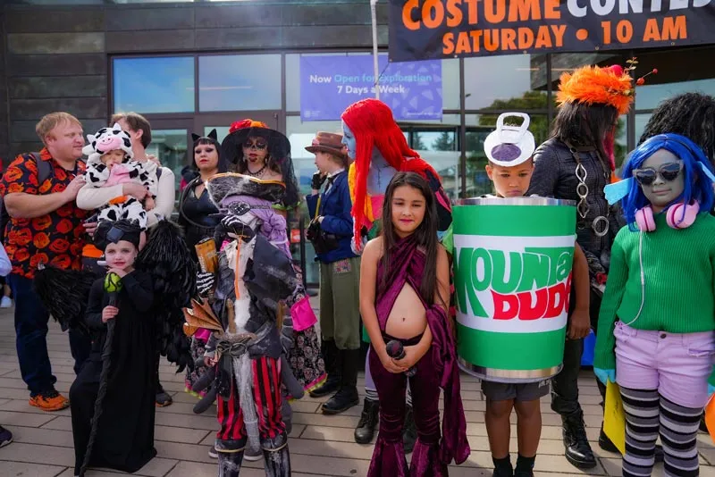 A group of people in various Halloween costumes, including a child dressed as a cow, a person in a cat costume, and a child dressed as a pirate, gather for a costume contest.