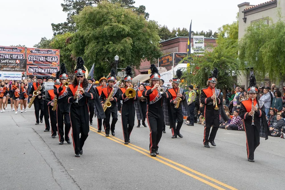 A marching band in uniform plays instruments as they parade down a street lined with spectators, with banners advertising the Half Moon Bay Pumpkin Festival in the background.