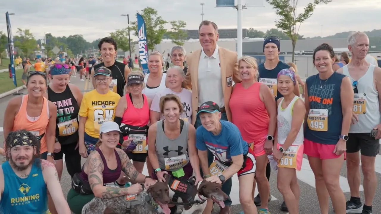 A diverse group of runners, some with dogs, pose for a photo with a man in a suit jacket at what appears to be the start line of a race.