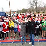 A large group of runners wearing Santa hats and festive costumes are gathered at the start line of a race, with two people in the foreground addressing the crowd.