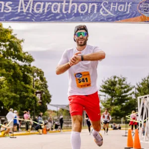 A male runner wearing a white t-shirt, red shorts, and sunglasses runs towards the camera under a race archway, checking his watch.