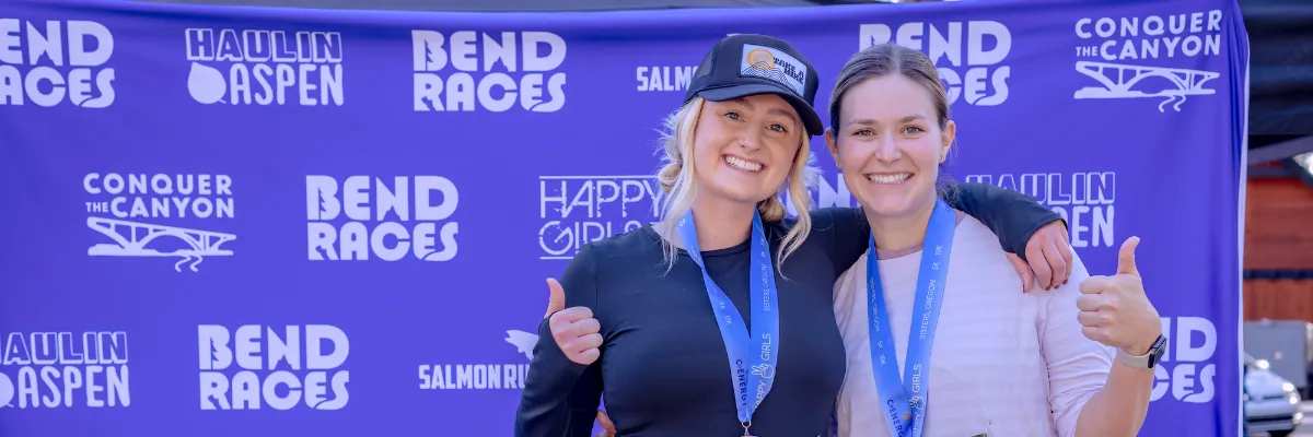 Two smiling women wearing race medals and giving thumbs up in front of a race banner.