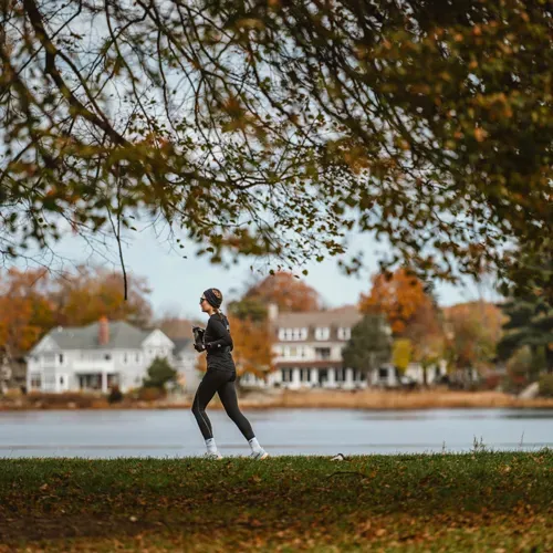 A woman runs along a path next to a lake, with houses and autumn trees in the background.