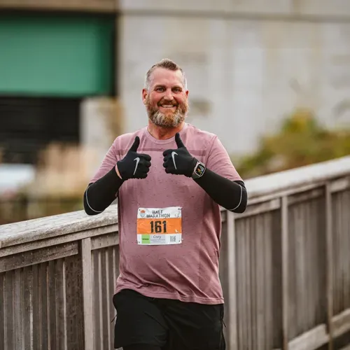 A male runner with a beard smiles and gives two thumbs up while wearing a race bib and running gear.