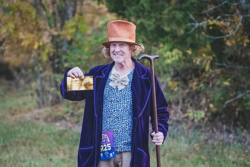 A man dressed as Willy Wonka, wearing a top hat and purple coat, holds up a golden ticket and a cane.