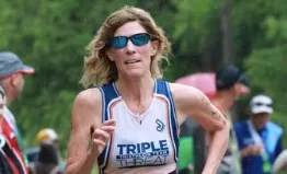 A female runner with sunglasses and a racing bib runs towards the camera.