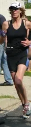 A female runner wearing a black tank top, black shorts, and a white baseball cap runs on a paved road.