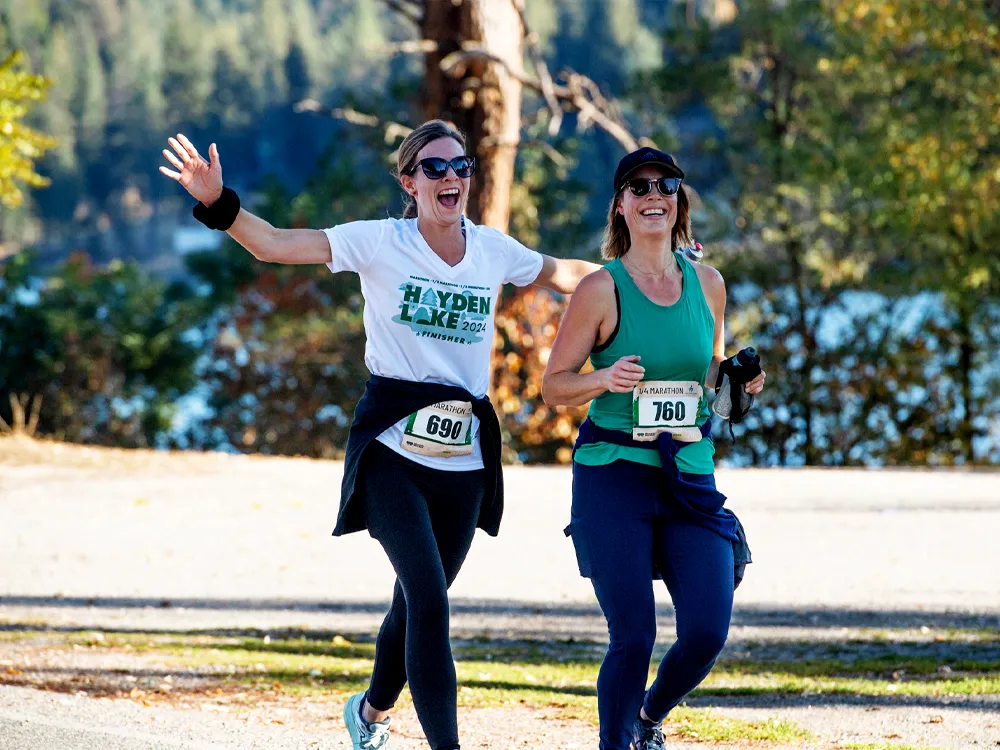 Two female runners smiling and running on a road with a lake in the background. One runner is raising her arm in celebration.