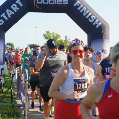 Runners gather at the start line under an inflatable archway, with a smiling woman in the foreground wearing red heart-shaped sunglasses.