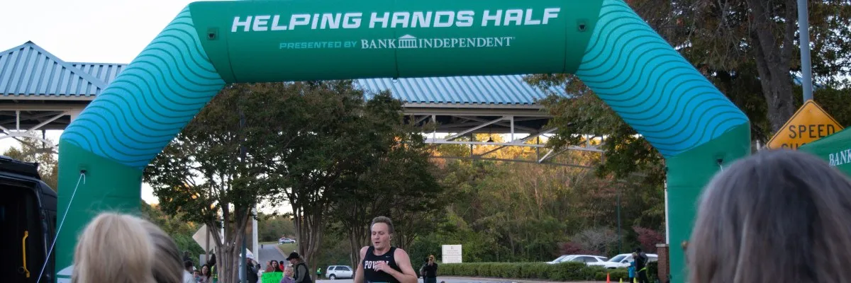 A male runner in a black tank top crosses under a green inflatable archway that reads "HELPING HANDS HALF".