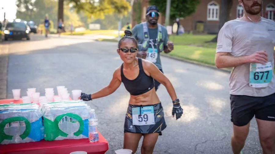 A female runner with her hair in a bun, wearing sunglasses and a black sports bra, reaches for a cup at a water station during a race. Other runners are visible in the background.