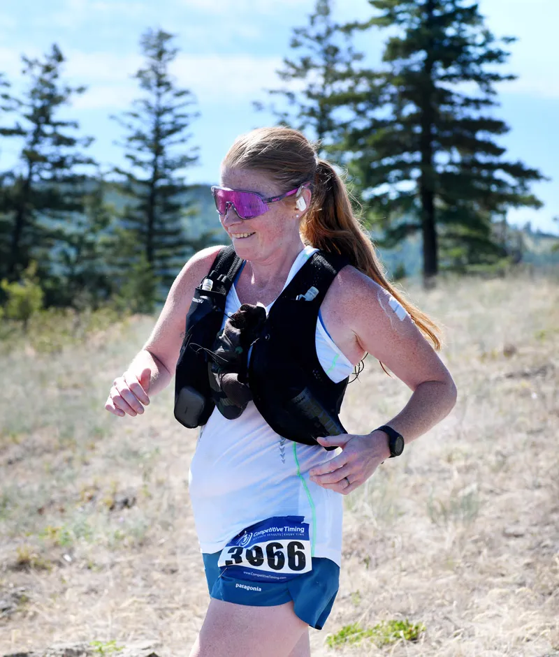 A female runner with red hair, wearing sunglasses and a hydration vest, runs on a dry, grassy trail with pine trees in the background.