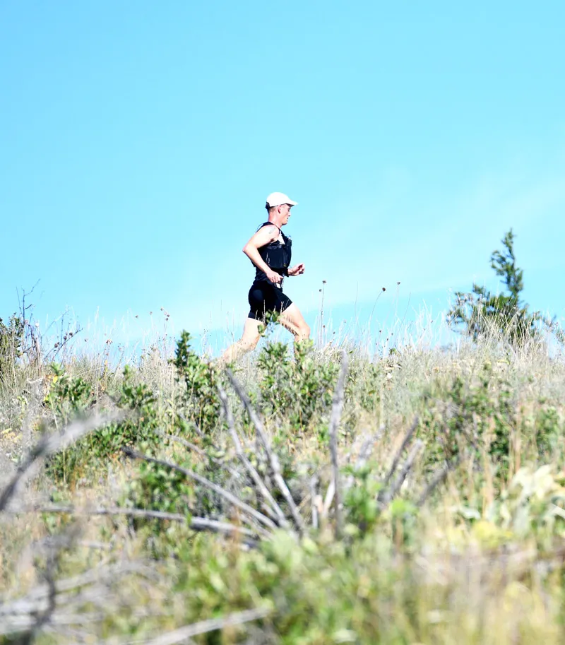 A runner in a black tank top and shorts runs uphill on a grassy, dry trail under a clear blue sky.