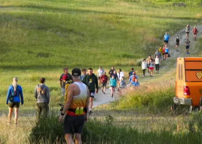 Runners on a dirt path through grassy hills, with an orange vehicle on the right side of the frame.