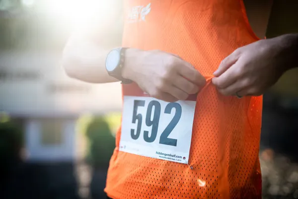 A runner fastens their bib number 592 onto an orange running vest, with a watch on their wrist.