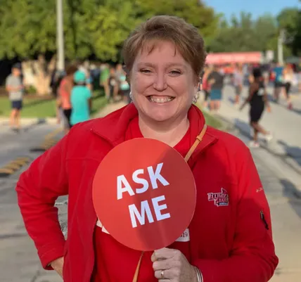 A smiling woman in a red jacket holds an 'ASK ME' sign at the start of a race, with runners in the background.