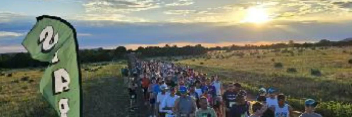 A large group of runners at the start line of a race, with a scenic sunset in the background and a green flag on the left.