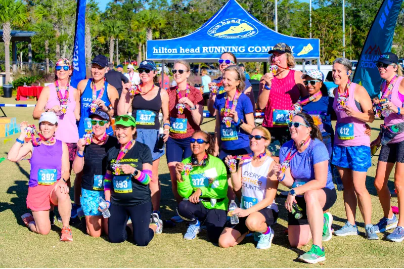 A group of female runners smile and show off their race medals in front of a blue tent.