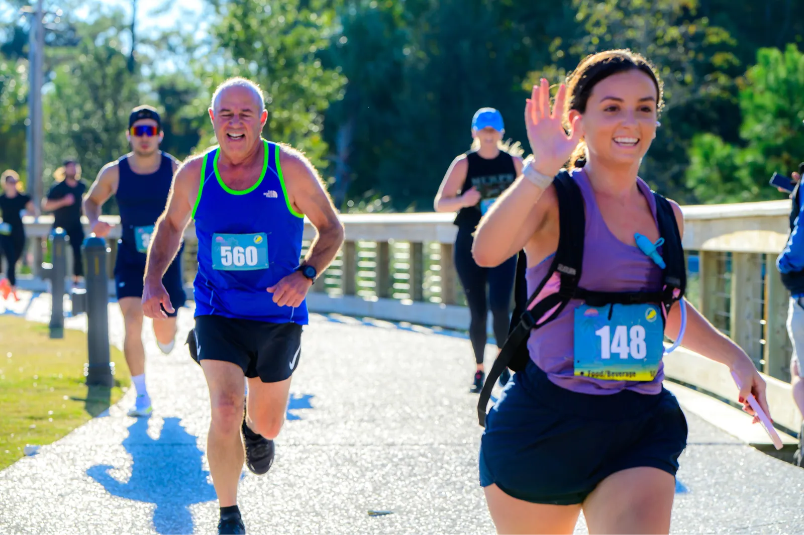Runners participating in a race, with one runner in the foreground waving and another runner in a blue singlet with bib number 560 smiling as they run.