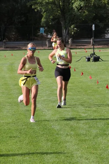 Two female runners in yellow tops and shorts running on a grassy field during a race.