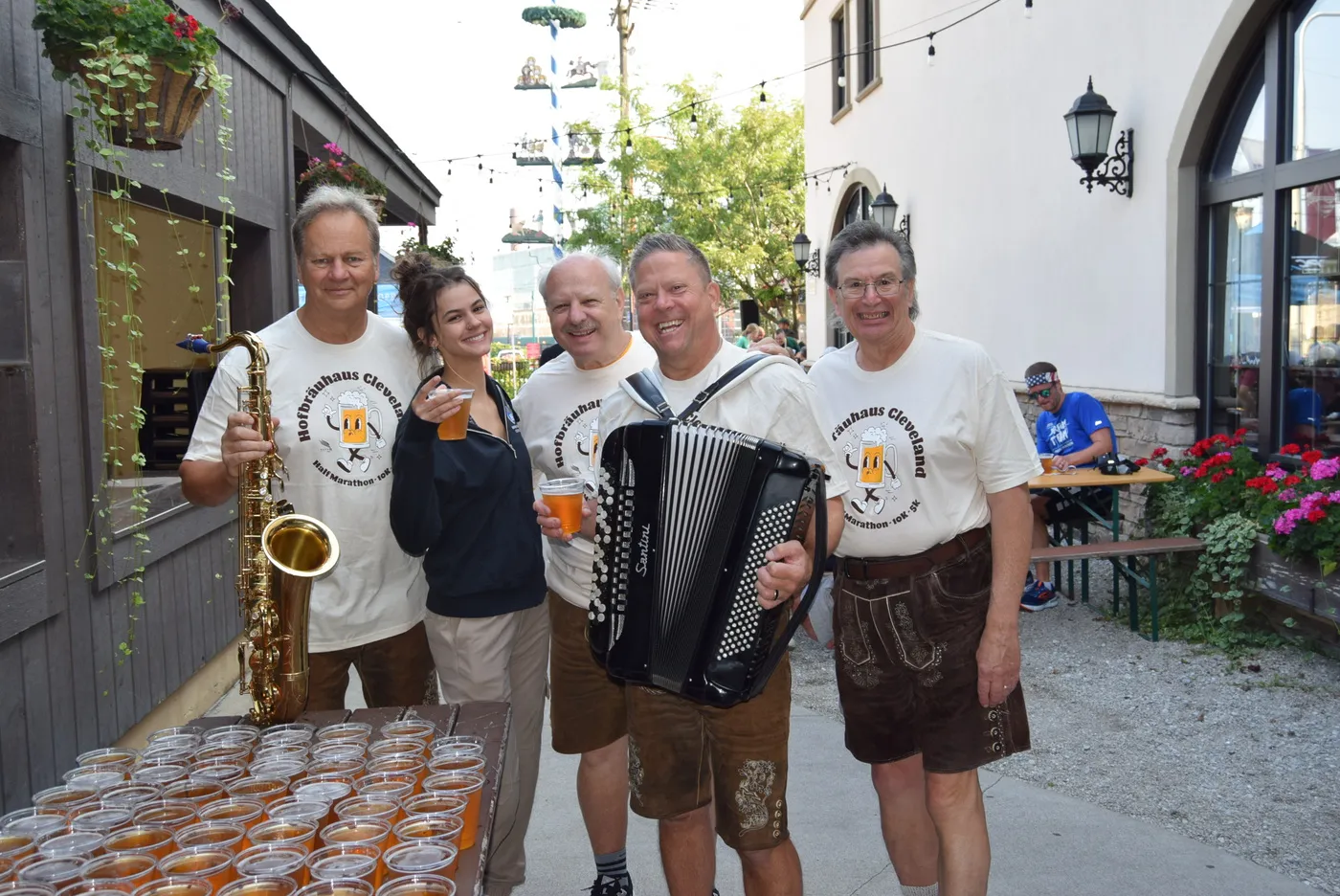A group of people, some holding musical instruments and drinks, wearing t-shirts that say 'Hofbräuhaus Cleveland Half Marathon 10K 5K'.