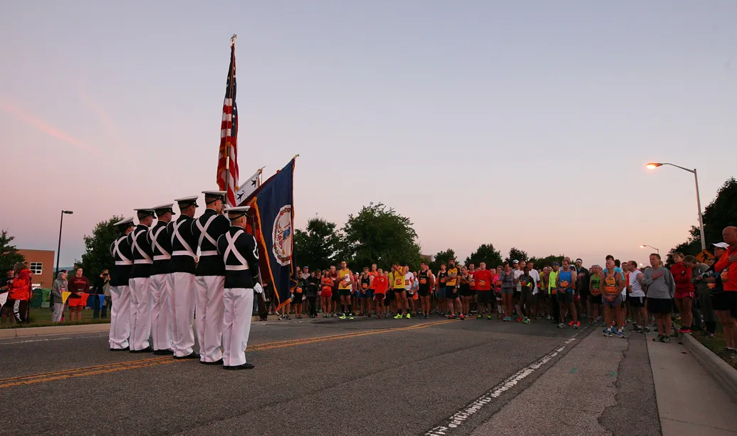 Runners gather at the start line of a race at dawn, with a color guard presenting flags.