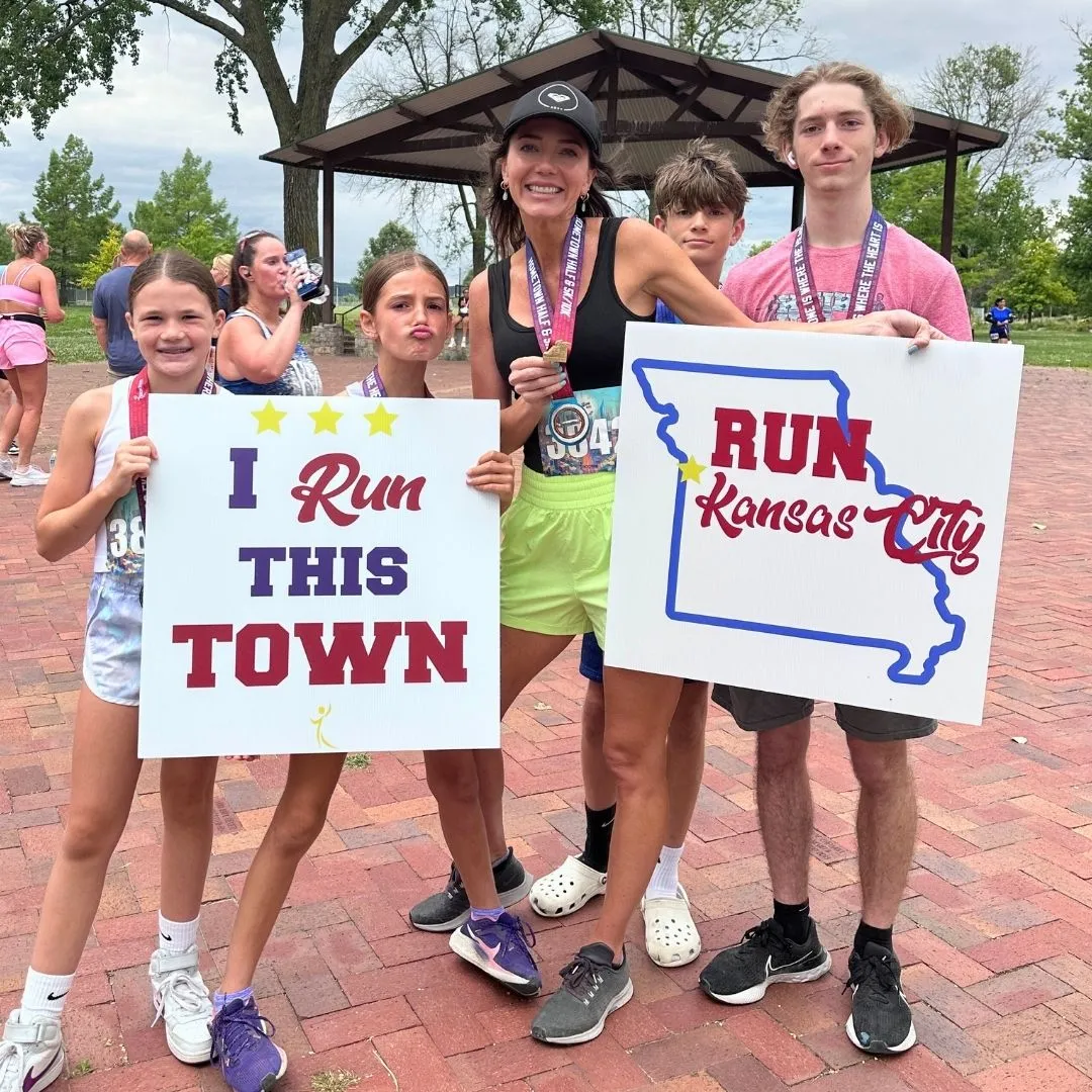 A group of people, including children and adults, pose for a photo after a race, holding signs that say "I Run This Town" and "RUN Kansas City".