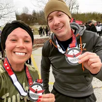 Two smiling runners, a woman and a man, pose for a selfie outdoors, both wearing race medals around their necks.