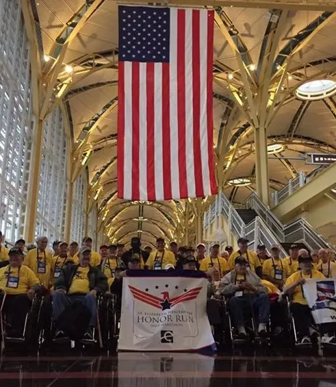 A group of people, including veterans in wheelchairs, stand together under a large American flag, holding a banner that reads 'Honor Run Half Marathon'.