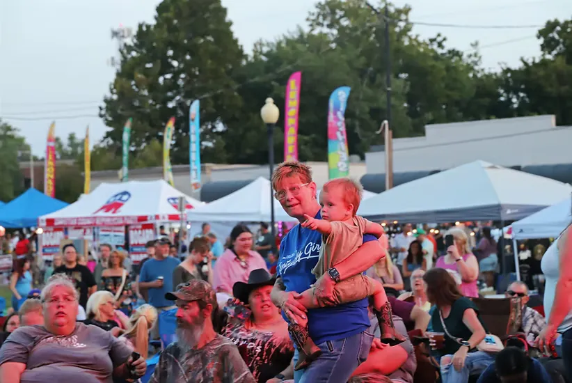 A woman holds a child while looking out at a crowd gathered at an outdoor event with tents and colorful flags in the background.