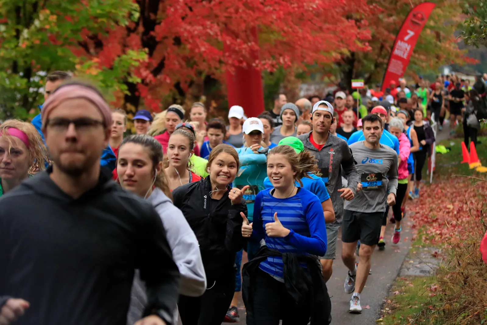 Runners smiling and giving thumbs up as they participate in a race, with colorful autumn trees in the background.