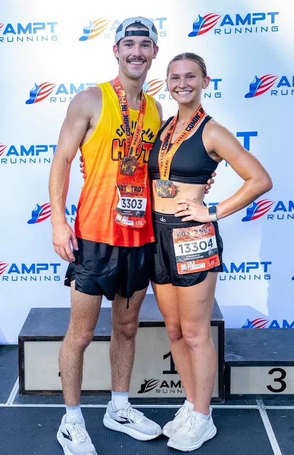 A male and female runner, both wearing medals and race bibs, smile at the camera while posing on a podium.