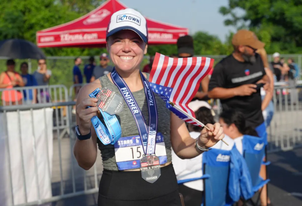 A smiling woman wearing a 'H-Town' hat and a race bib holds up two finisher medals and a small American flag.