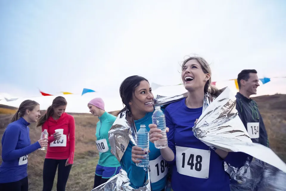 Two women wrapped in silver Mylar blankets smile and laugh after finishing a race, holding water bottles.