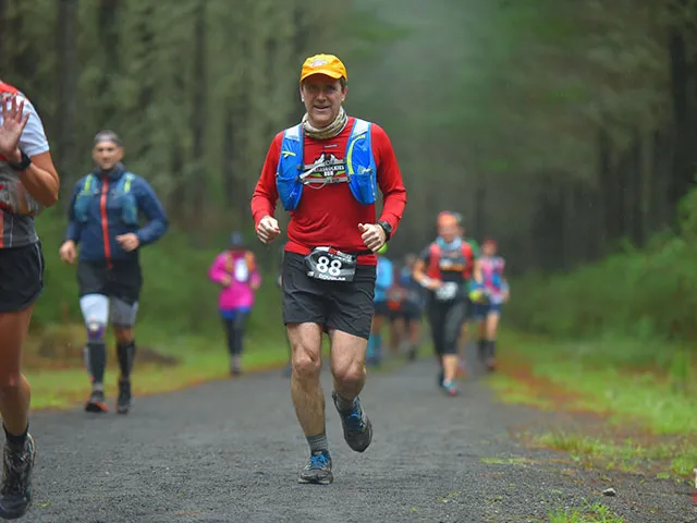 A male runner in a red shirt and yellow hat smiles as he runs on a gravel path through a forest, with other runners behind him.