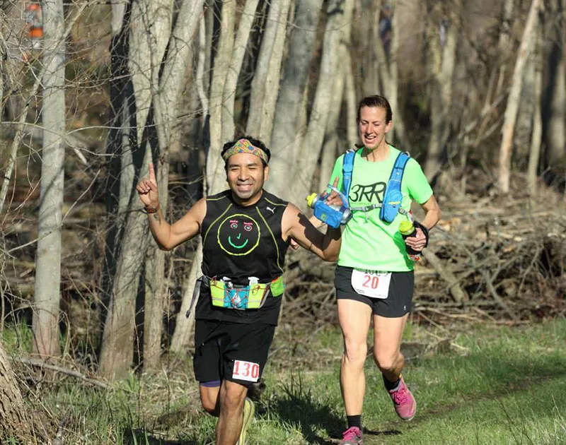 Two runners, one with a smiley face on his shirt and a headband, and another in a bright green shirt, running on a wooded trail.