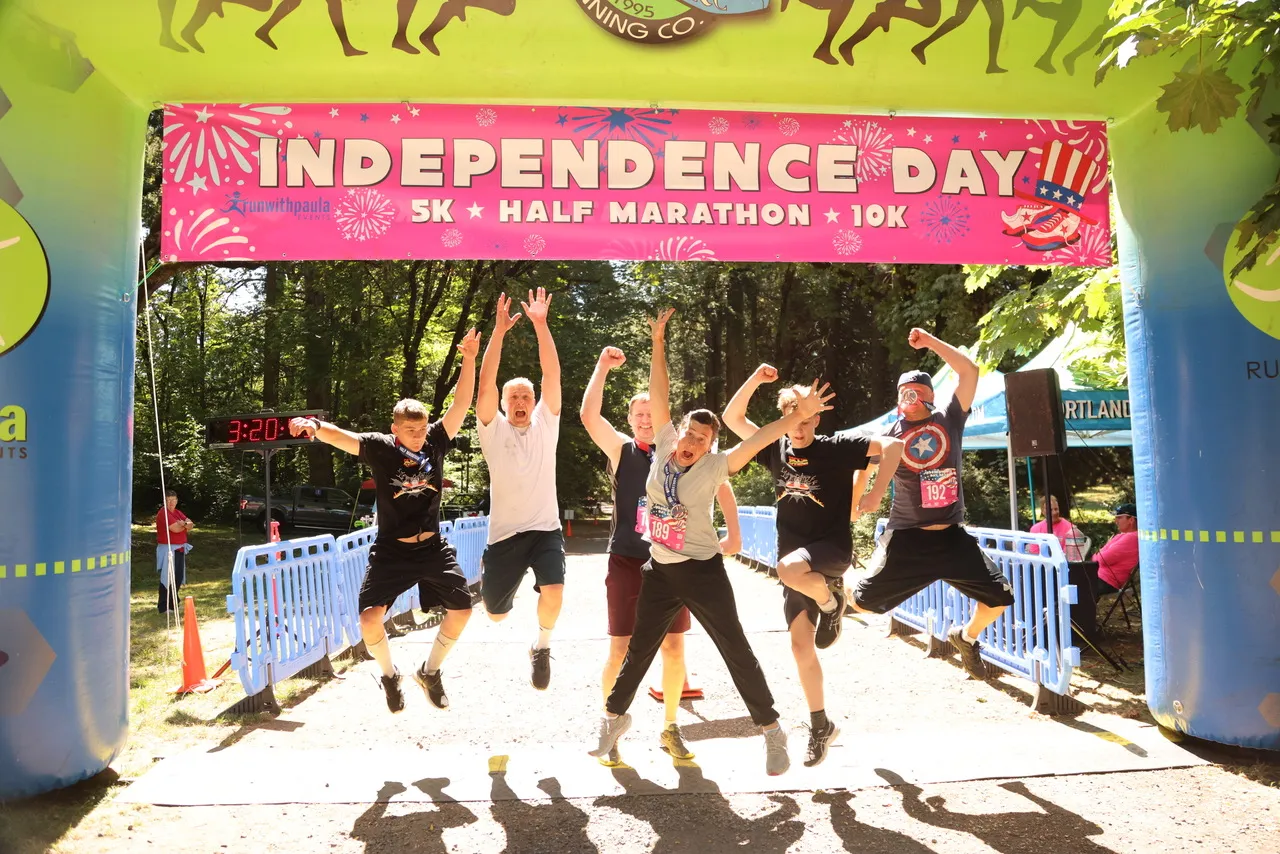 A group of runners joyfully jumping in the air under the Independence Day Half Marathon start/finish line arch.