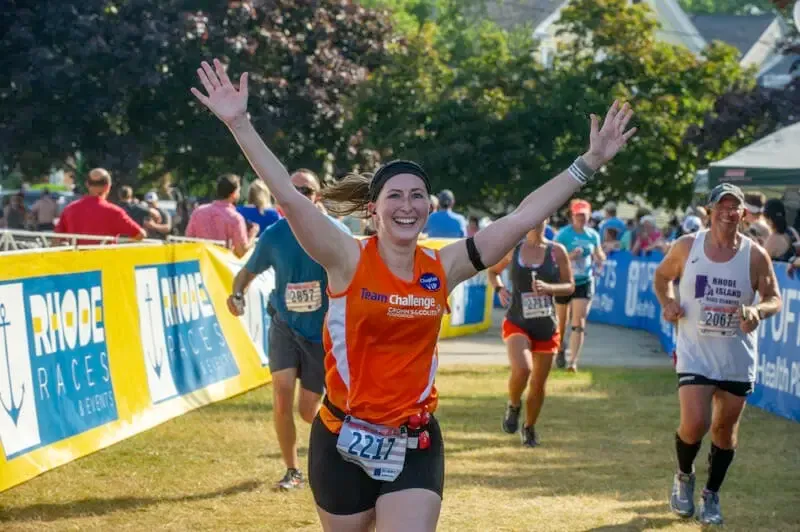 A female runner with her arms raised in celebration crosses the finish line of the Independence Rhode Race, smiling broadly. Other runners and race banners are visible in the background.