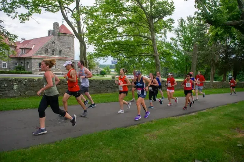 A group of runners on a paved path with a stone building and trees in the background.