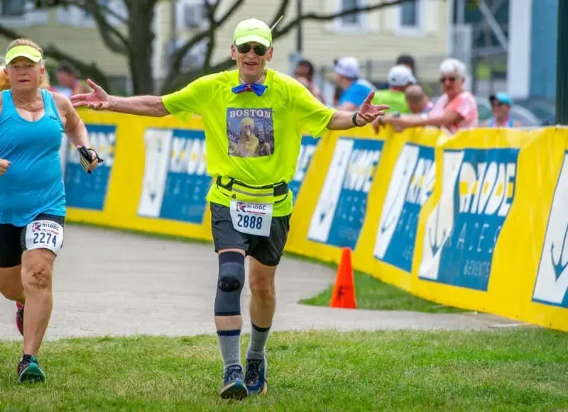 A male runner with arms outstretched, wearing a bright yellow shirt and bib number 2888, runs towards the camera with a female runner behind him.