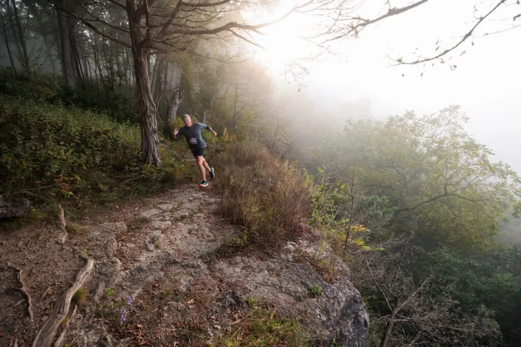 A male runner navigates a foggy, wooded trail with rocky terrain.