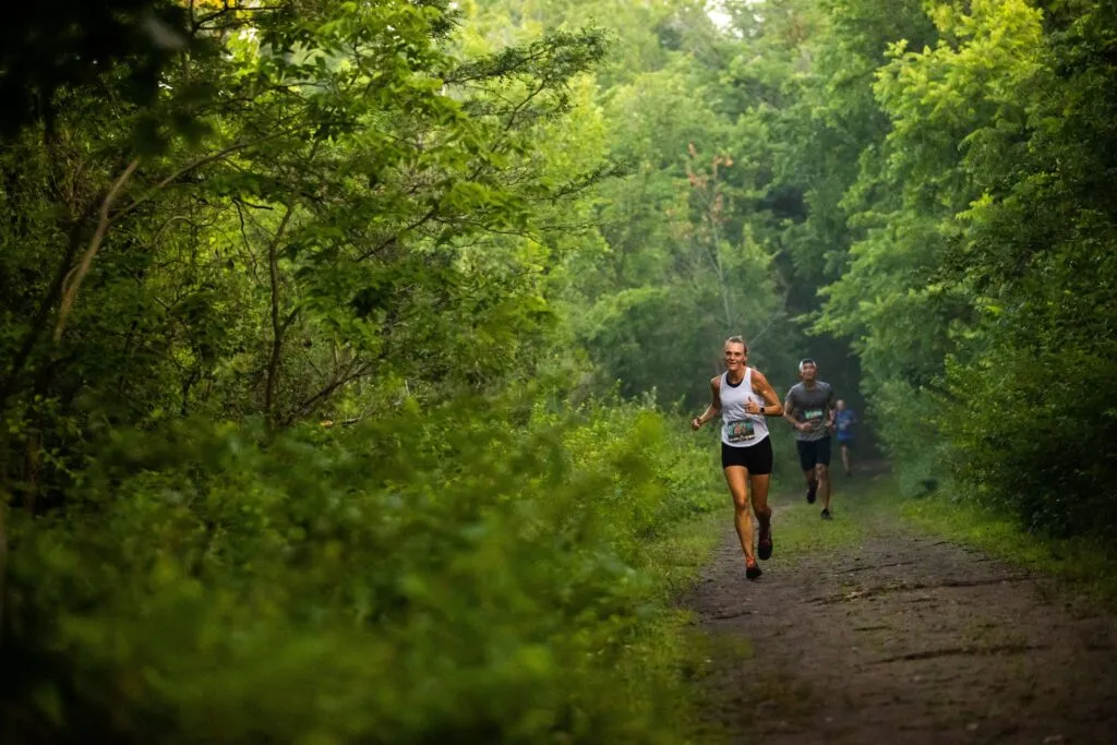 A female runner smiles as she runs down a dirt trail surrounded by lush green forest, with other runners behind her.