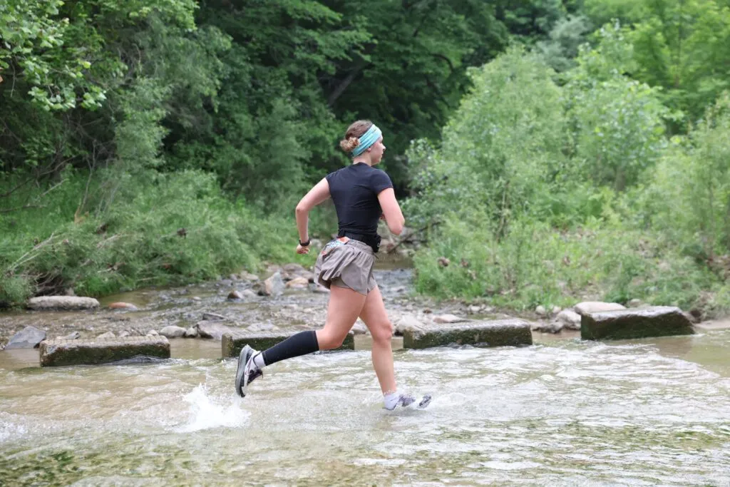 A female runner splashes through a shallow stream on a wooded trail.