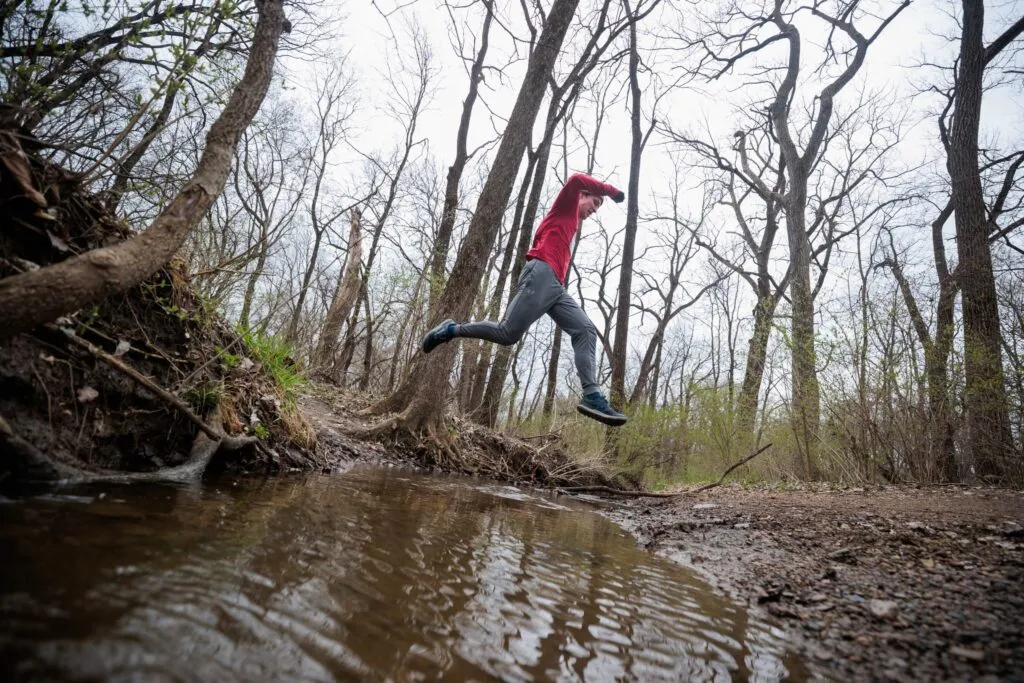 A runner in a red long-sleeved shirt and grey pants jumps over a small stream on a wooded trail.