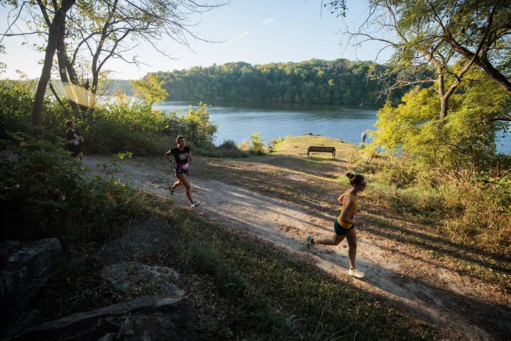 Runners on a dirt path next to a lake with trees in the background.