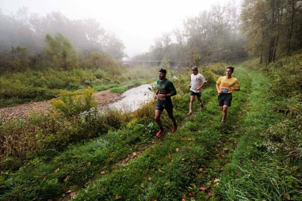 Three runners on a foggy trail next to a creek, surrounded by lush green foliage and trees.