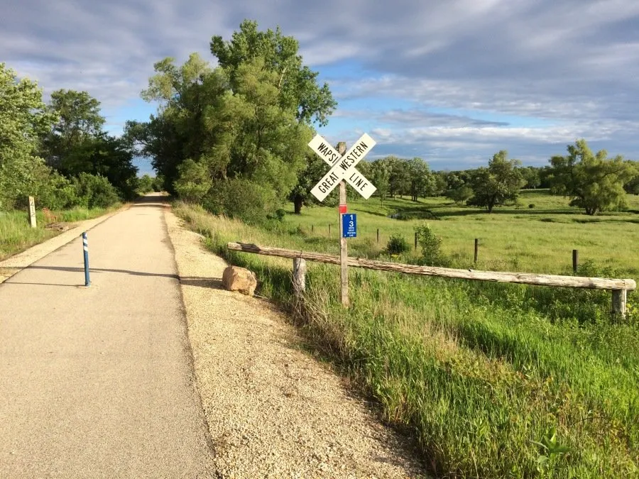 A paved trail runs through a scenic landscape with trees and a railroad crossing sign.
