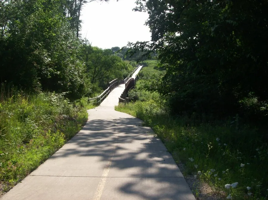 A paved path curves towards a wooden bridge over a grassy area, surrounded by trees.