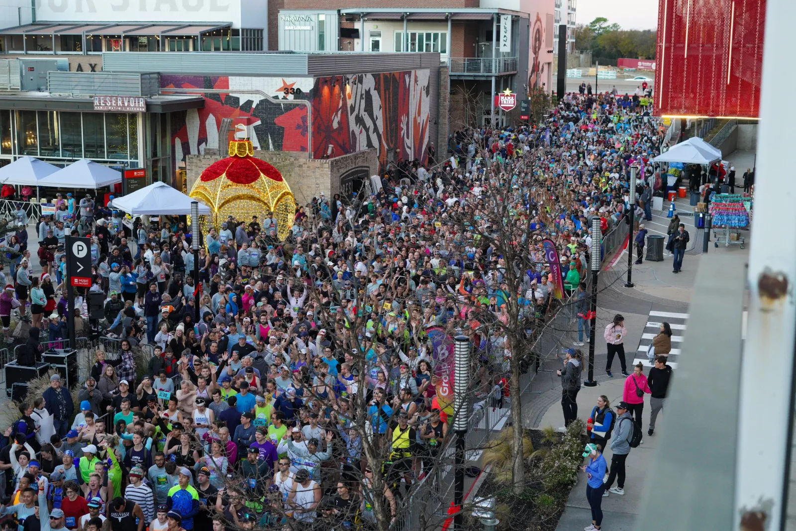 A large crowd of runners gathered at the start line of a race, with many people waving and looking towards the camera. The urban setting features festive decorations, including a large illuminated ornament.