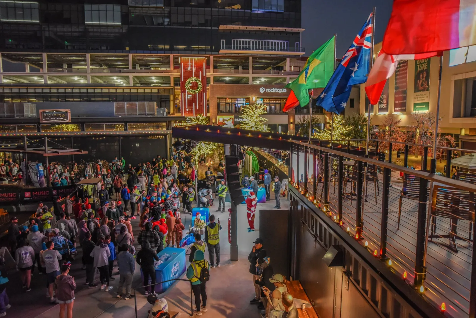 A lively crowd of people gathered at an outdoor event, with festive lights and decorations, possibly at the start or finish line of a race.
